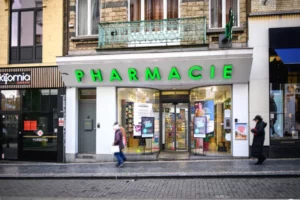Façade d'une pharmacie à Bruxelles avec passants, enseigne lumineuse et affiches de produits pharmaceutiques.